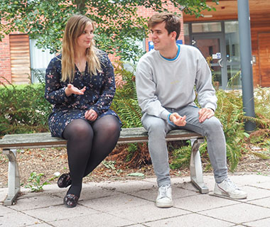 two students are talking whilst sitting on a bench